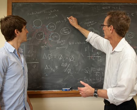 A professor gestures toward diagrams on a chalkboard as student looks on.